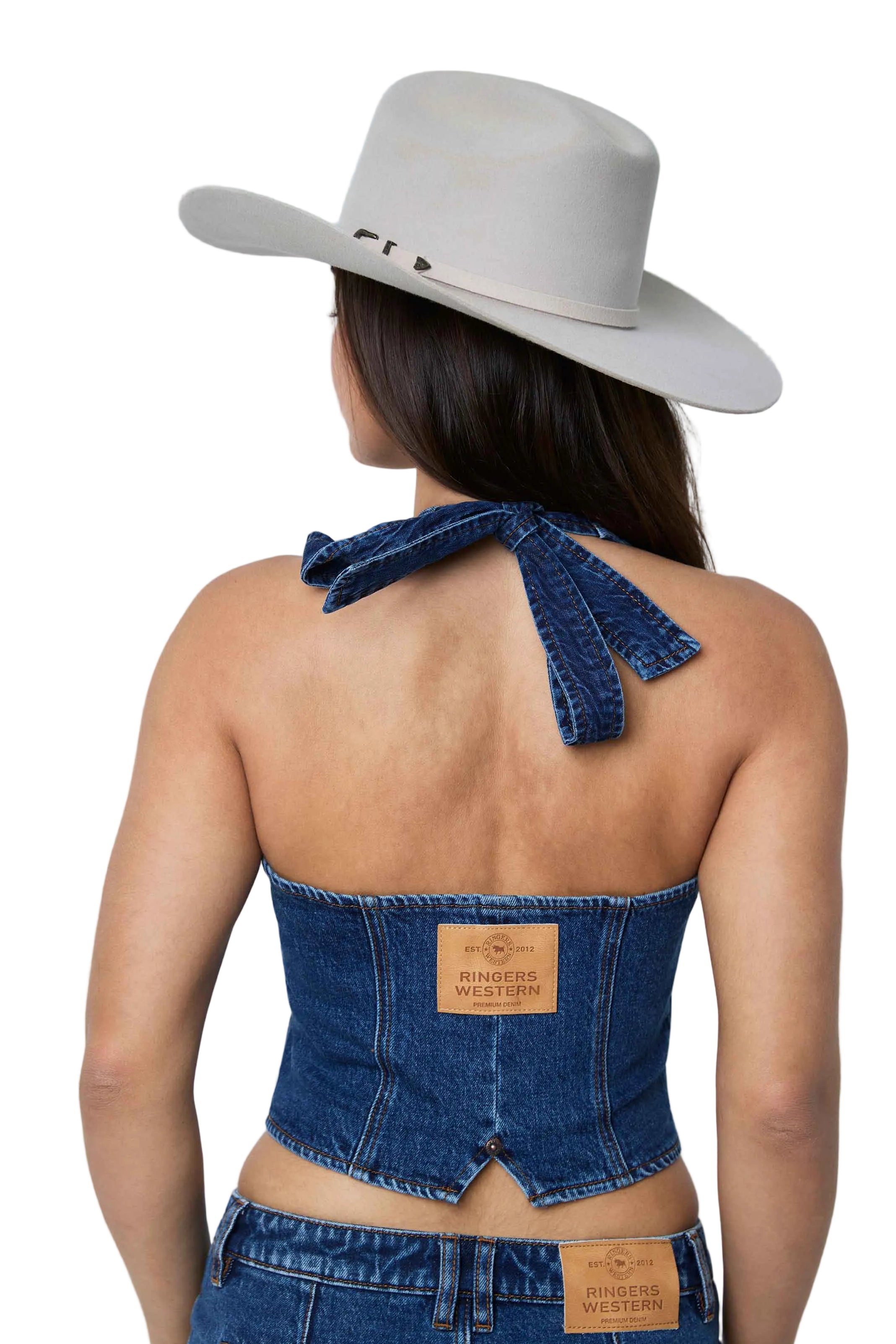 A woman shown from the back wearing a blue denim halter-style corset top with a tie at the neck, paired with a light beige cowboy hat and denim bottoms.