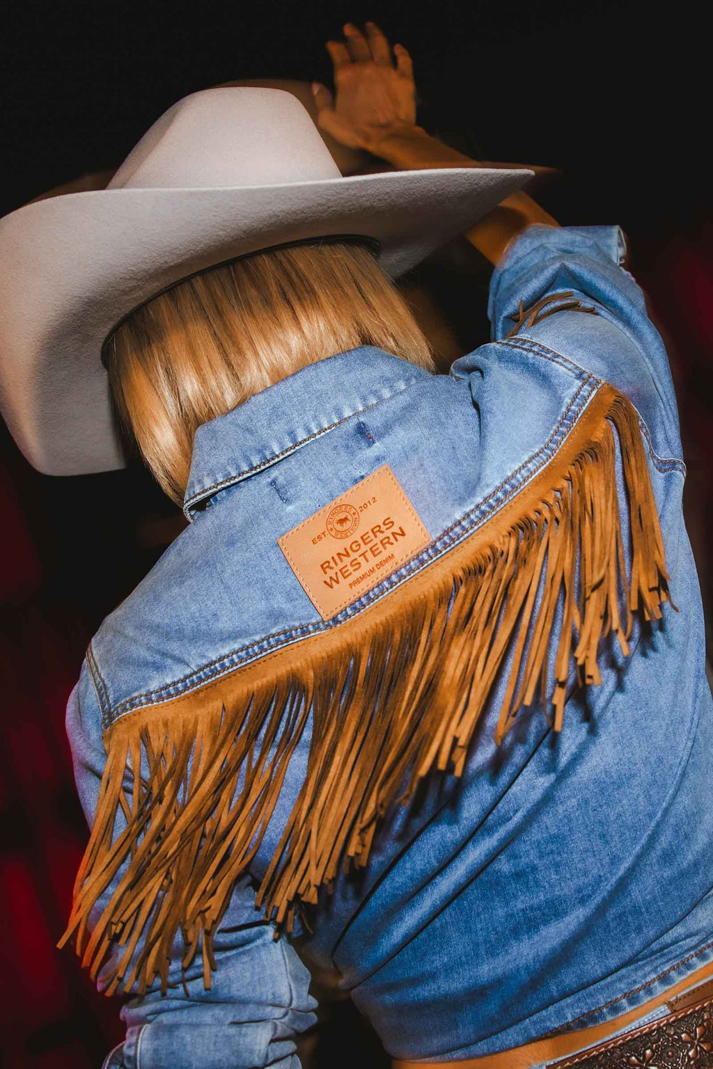A close-up back view of a person wearing a light blue denim jacket with a large tan fringe across the shoulders and a tan leather patch on the upper back that reads 'RIDGE WESTERN'. The person is also wearing a white cowboy hat, and the background is very dark.