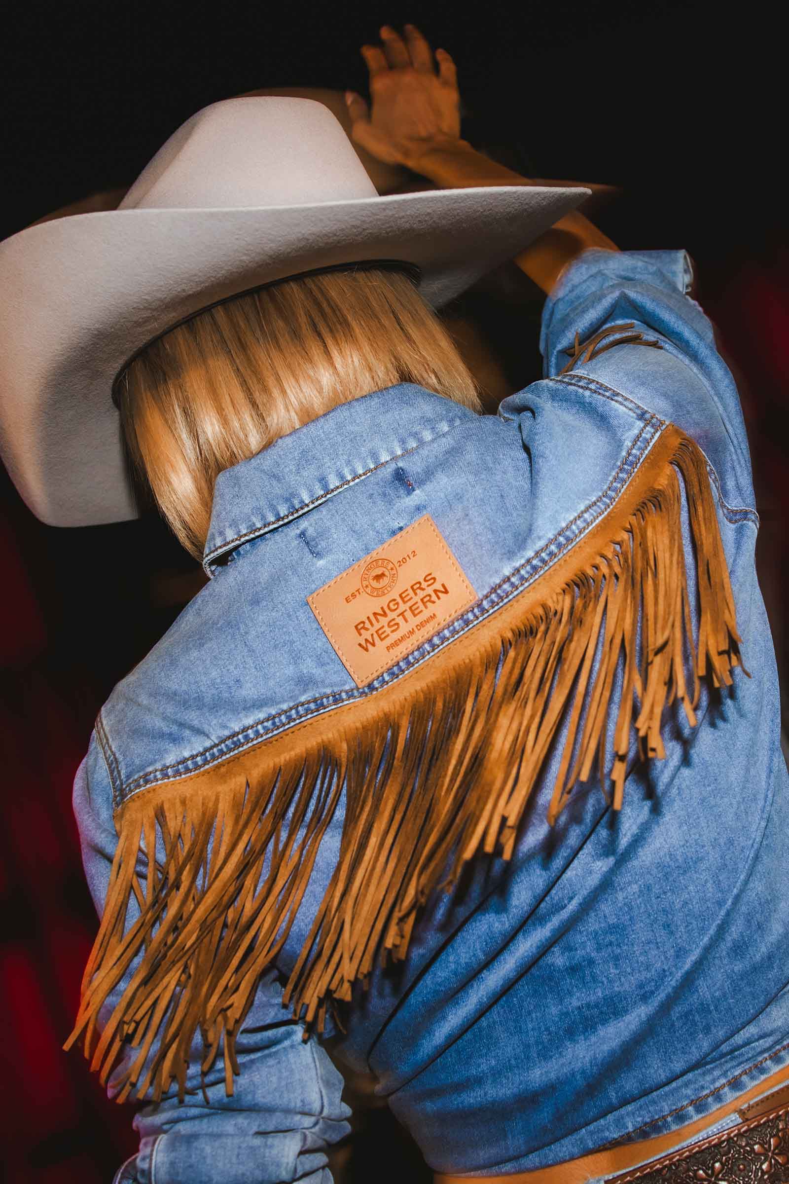 A close-up back view of a person wearing a light blue denim jacket with a large tan fringe across the shoulders and a tan leather patch on the upper back that reads 'RIDGE WESTERN'. The person is also wearing a white cowboy hat, and the background is very dark.