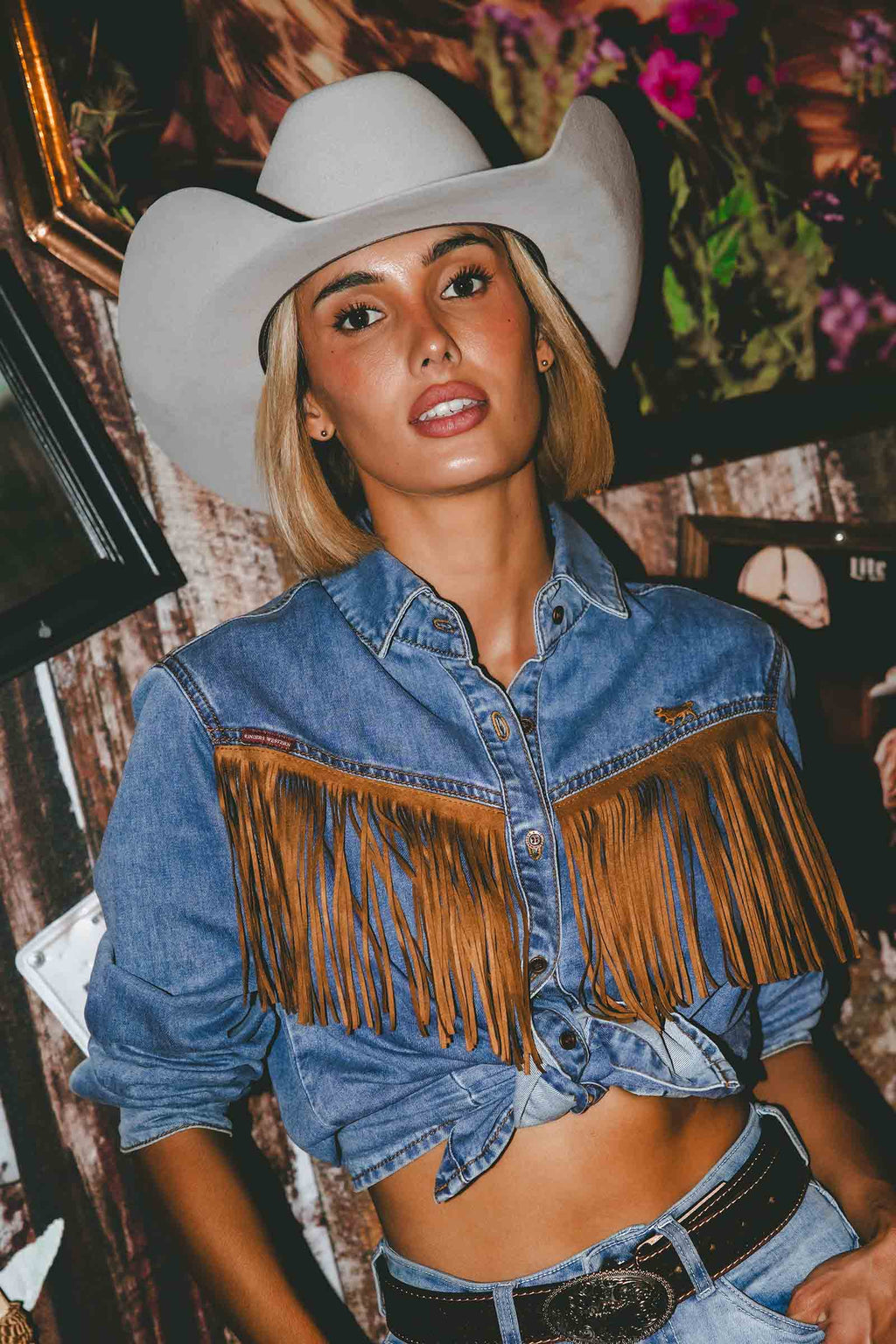 A woman wearing a light gray cowboy hat and a cropped blue denim shirt with large brown fringe across the chest, paired with a black belt, posed in a decorated indoor setting.