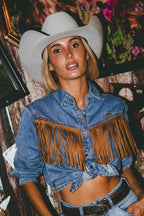A woman wearing a light gray cowboy hat and a cropped blue denim shirt with large brown fringe across the chest, paired with a black belt, posed in a decorated indoor setting.