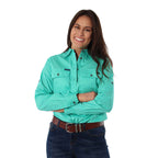 A woman with long dark hair smiles with her arms crossed, wearing a Ringers Western Womens Pentecost River Half Button Work Shirt in Mint and blue jeans with a brown belt. The background is plain white.