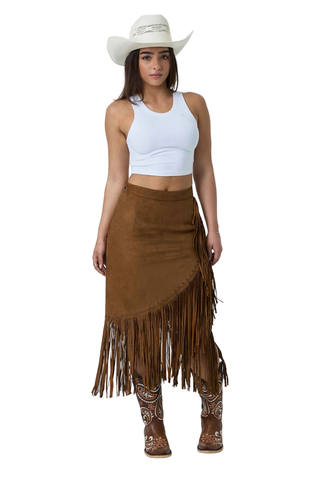 A woman modeling a brown suede fringe skirt with a white crop top, a white cowboy hat, and brown western boots against a white studio background.