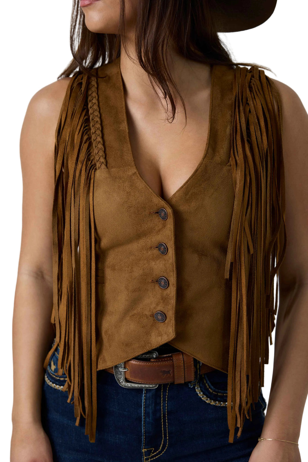 A woman wearing a brown suede fringe vest with a button-front closure and braided shoulder detailing, paired with blue jeans and a dark hat against a white studio background.
