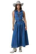 A woman modeling a sleeveless blue denim midi dress with a button-front bodice, tie closure, and gathered skirt, styled with a light beige cowboy hat and white cowboy boots against a white studio background.