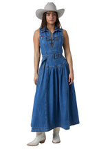 A woman modeling a sleeveless blue denim midi dress with a button-front bodice, tie closure, and gathered skirt, styled with a light beige cowboy hat and white cowboy boots against a white studio background.
