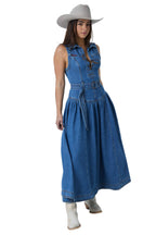 A woman modeling a sleeveless blue denim midi dress with a button-up collar, tie waist, and flared skirt, styled with a light beige cowboy hat and white boots against a white studio background.