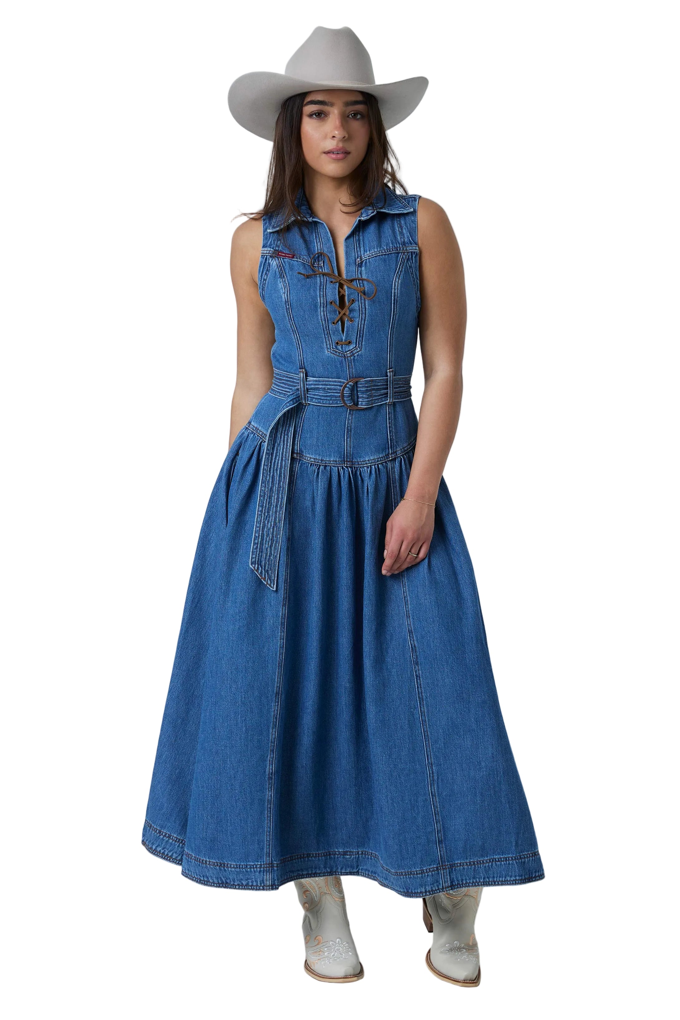 A woman modeling a sleeveless blue denim midi dress with a tie-front detail, matching belt, and flared skirt, styled with a light gray cowboy hat and white cowboy boots against a white studio background.