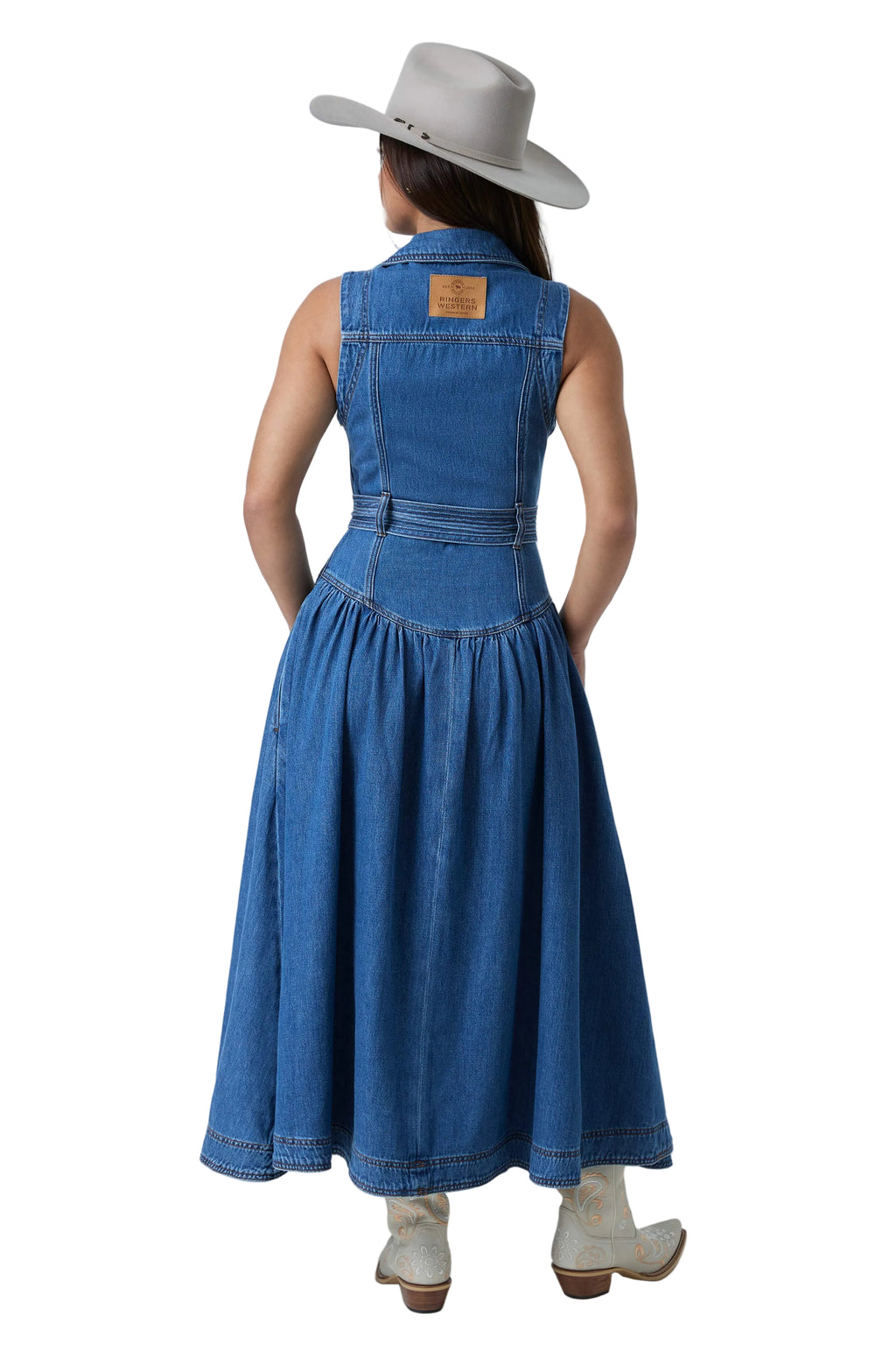 A woman shown from the back wearing a sleeveless blue denim midi dress with a gathered waist and flared skirt, paired with a light gray wide-brim hat and white cowboy boots, against a white studio background.