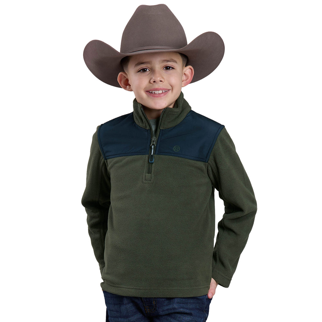 A young boy in a Roper Boys L/S Pullover (39692209) Green by Roper, featuring an olive green color with a dark green yoke and reflective zipper pulls, smiles with his hands in his pockets against a white background.