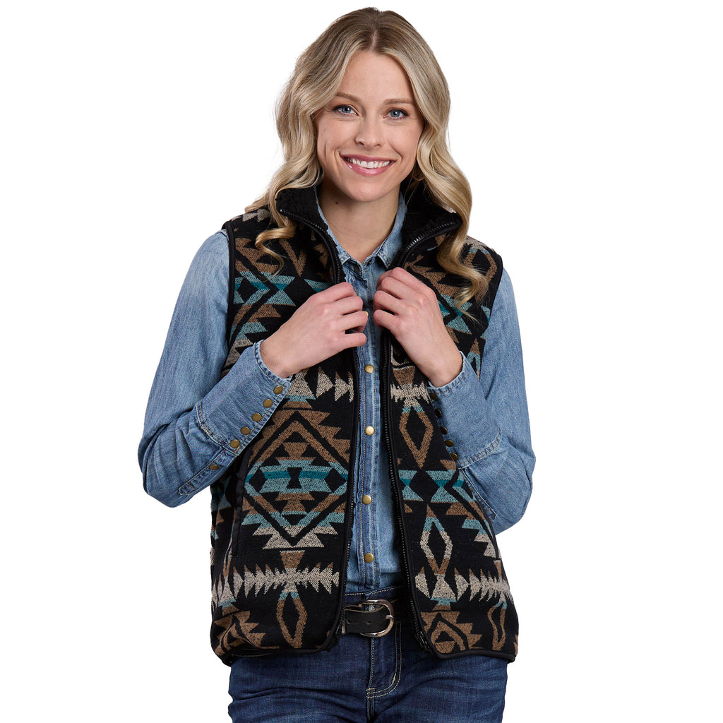 A smiling woman with wavy blonde hair wears a Roper Womens Vest (98781128) Black over a blue denim shirt, holding the vest's collar with both hands against a white background. The vest features black, turquoise, and brown geometric designs.