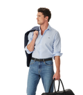 A man in an R.M.Williams Mens Sommers Poplin Stripe L/S Shirt (SH200PM01) White Blue, blue jeans, and a black belt holds a dark jacket over his shoulder and carries a black duffel bag, looking left against a white background.
