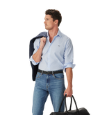 A man in an R.M.Williams Mens Sommers Poplin Stripe L/S Shirt (SH200PM01) White Blue, blue jeans, and a black belt holds a dark jacket over his shoulder and carries a black duffel bag, looking left against a white background.