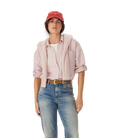 A woman in an R.M.Williams Womens Hopeland Linen Check L/S Shirt (Ice Pink), white top, blue jeans with a brown belt, and a red cap stands against a plain white background.