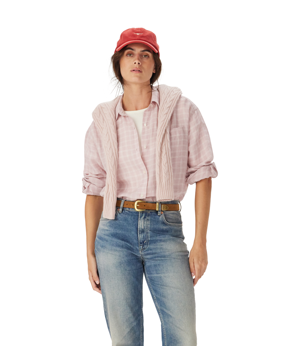 A woman in an R.M.Williams Womens Hopeland Linen Check L/S Shirt (Ice Pink), white top, blue jeans with a brown belt, and a red cap stands against a plain white background.
