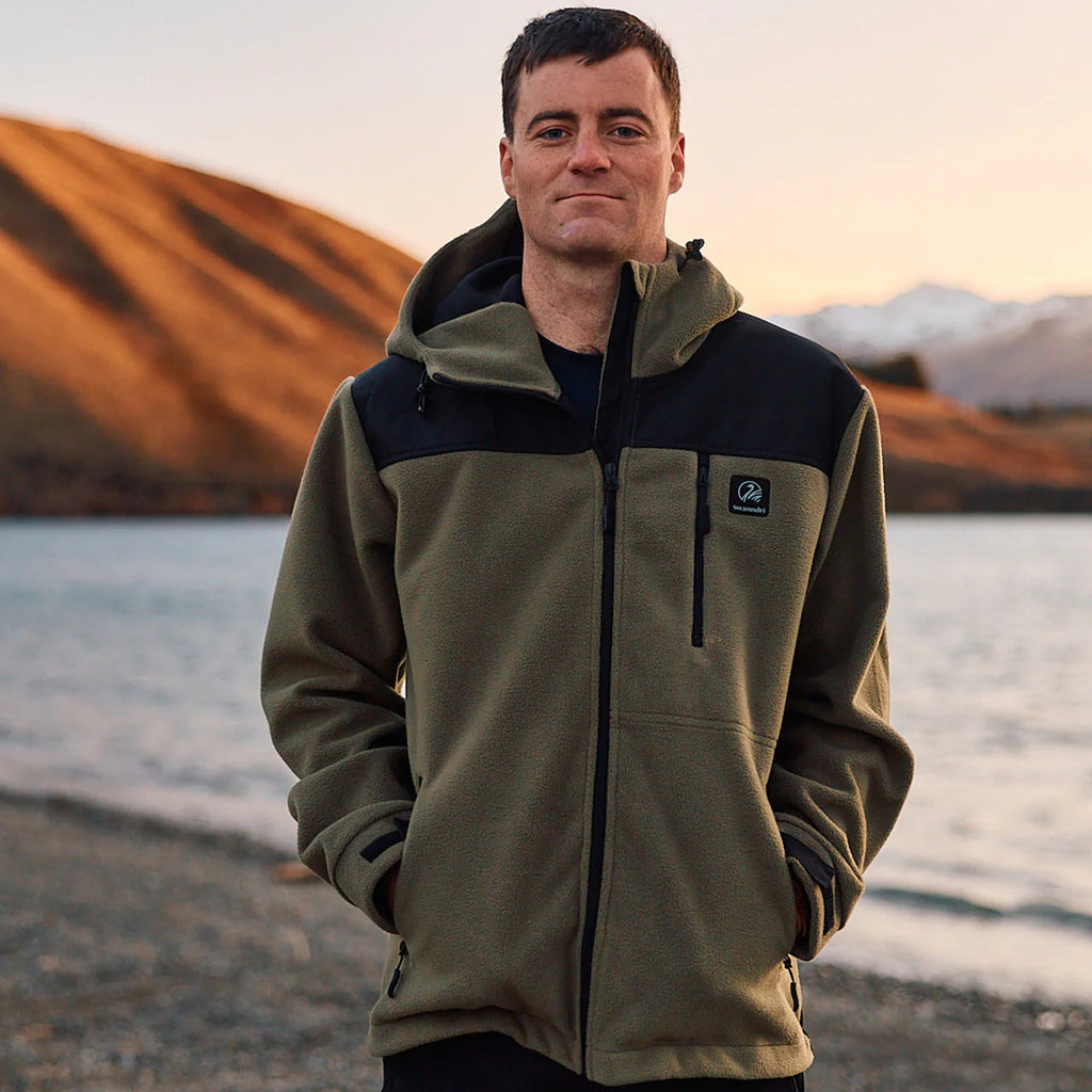 A man stands by a lake, wearing the Swanndri Men's High Rock Stormshield Jacket (SW2562070M) in Forest, an olive green weatherproof fleece with black yoke and lower back