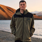 A man stands by a lake, wearing the Swanndri Men's High Rock Stormshield Jacket (SW2562070M) in Forest, an olive green weatherproof fleece with black yoke and lower back