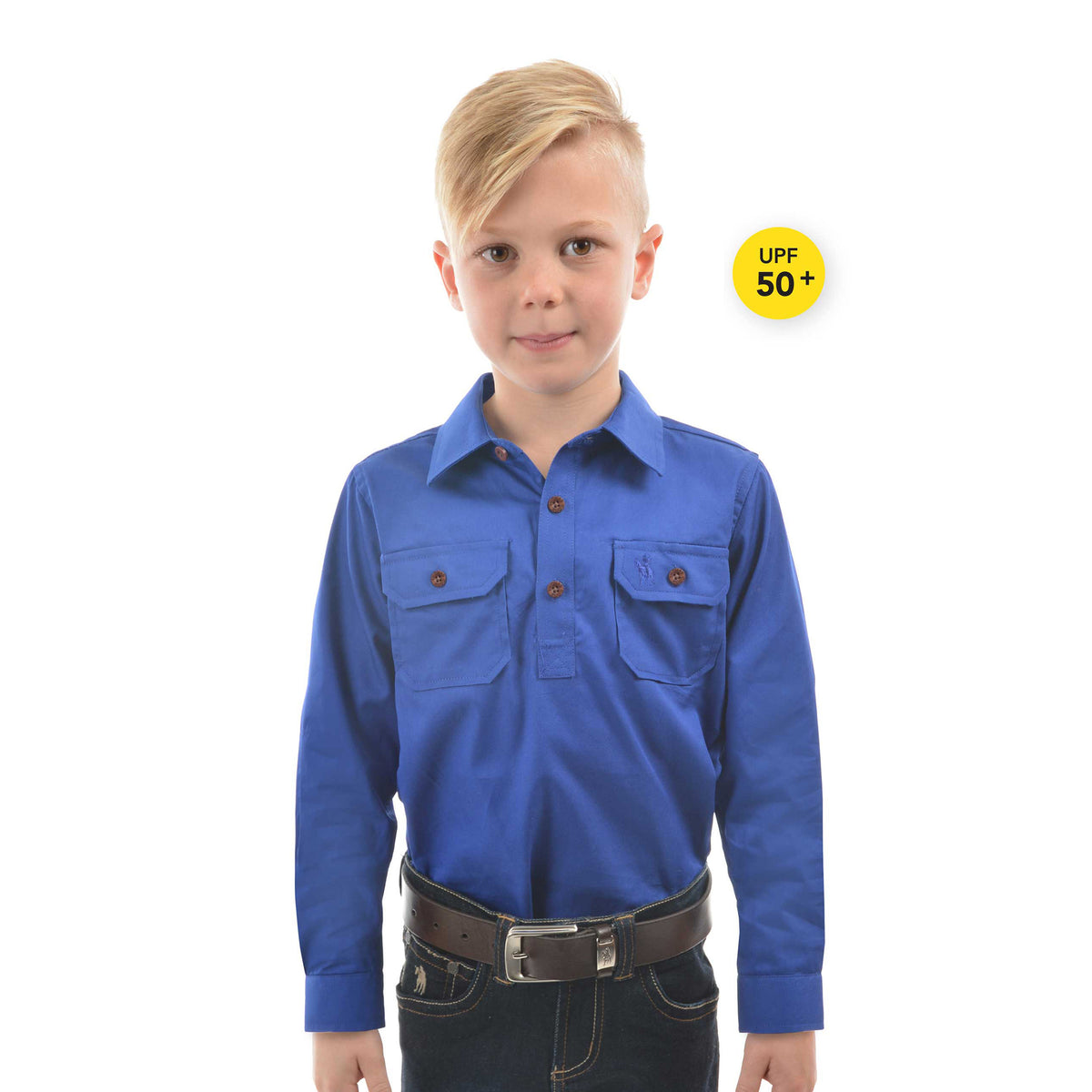 A young boy modelling a cobalt blue Thomas Cook Children's Heavy Drill Half Button Long Sleeve Shirt (TCP7100163) with two chest pockets, paired with dark jeans and a brown belt, on a white background with a yellow 'UPF 50+' badge.