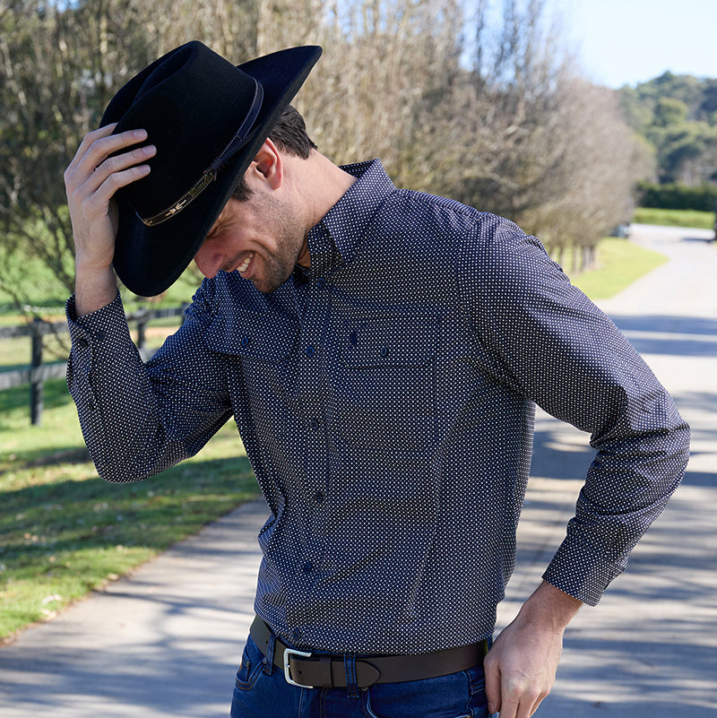 A man wearing a dark navy long-sleeve button-down shirt, blue jeans, and a black Thomas Cook Kimberley Crushable Hat (TCP1937002), standing on a road outdoors.