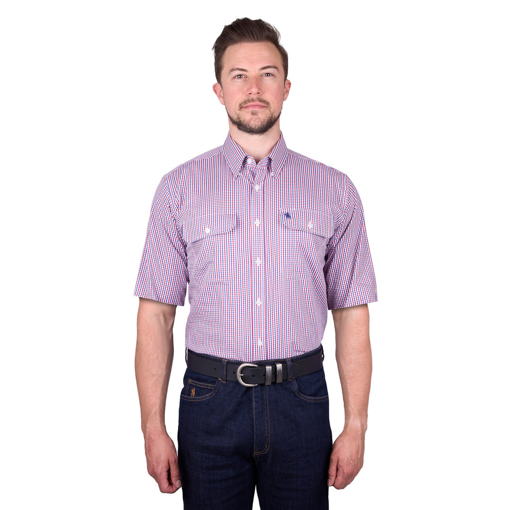 A man wearing a royal blue, white and red Thomas Cook Men's Wentworth Short Sleeve Dress Shirt (TCP1110158) with two chest pockets, paired with dark blue jeans and a black belt, against a plain white background.