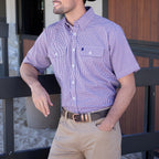 A man wearing a royal blue, white and red Thomas Cook Men's Wentworth Short Sleeve Dress Shirt (TCP1110158) with two chest pockets, paired with camel coloured moleskin pants and a brown belt, leaning against a black railing.