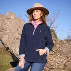 A woman wearing a navy quarter-zip Thomas Cook Women's Charlie Classic 1/4 Zip Neck Rugby (TCP2511030) with a pink zipper and a pink, white, and blue striped inner collar, denim jeans and a tan wide-brim hat, posing outdoors against a large tree trunk under a clear blue sky.