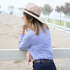 A woman wearing a beige wide-brim hat with a brown band, a blue and white striped Thomas Cook Women's Lawson Long Sleeve Shirt (TCP2112178) with vertical blue and white stripes, paired with dark pants and a belt, a leans on a white fence outdoors at a ranch.
