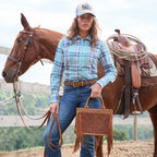 A woman stands outdoors beside a brown horse, wearing a teal and pink plaid Wrangler Women Luisa Long Sleeved Shirt (X6S2100607), blue Wrangler Etta High Rise Bootcut Jeans Bailey (XCP2255711) with a tan Wrangler Siara Belt (X6S2916BLT) and a Wrangler Josie High Profile Trucker Cap X6S2943CAP, while holding a tan Wrangler Adelyn Bag (X6S2975BAG) fringe handbag. The horse has a saddle and lariat on it, with a fence and trees in the background.