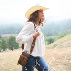 A woman wearing a white embroidered Wrangler Ava Blouse (X6S2501614), mid indigo Wrangler Etta Bootcut Jeans Bailey (XCP2255711), and a Jhonson Bangora Western Hat (X4Y2025JHO) walks outdoors in a grassy field while carrying a tan leather Camile Wallet Bag (X6S2977BAG) with a tan strap.