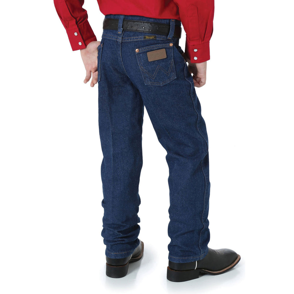 A boy shown from behind, from the waist down wearing a pair of prewashed indigo Wrangler Boys Junior Original ProRodeo Regular Fit Jeans (13MWZJPREG) paired with western boots, a western belt and a red shirt on a white background.