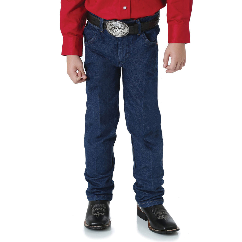 A boy shown from the waist down wearing a pair of prewashed indigo Wrangler Boys Junior Original ProRodeo Regular Fit Jeans (13MWZJPREG) paired with western boots, a western belt and a red shirt on a white background.