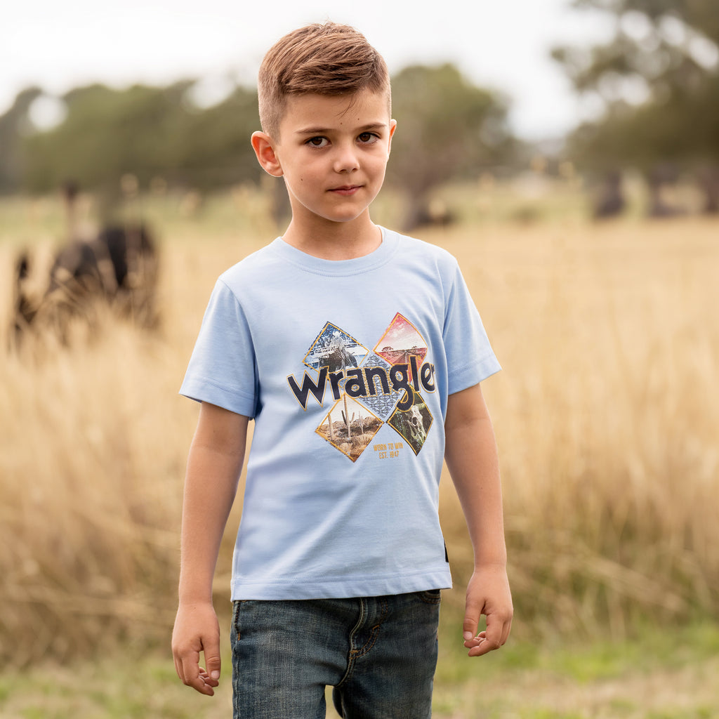 A young boy wearing a light blue Wrangler Boys Mark Short Sleeve Tee (X6S3557688) with a graphic print on the front, paired with dark blue jeans, photographed standing in a grassy field.