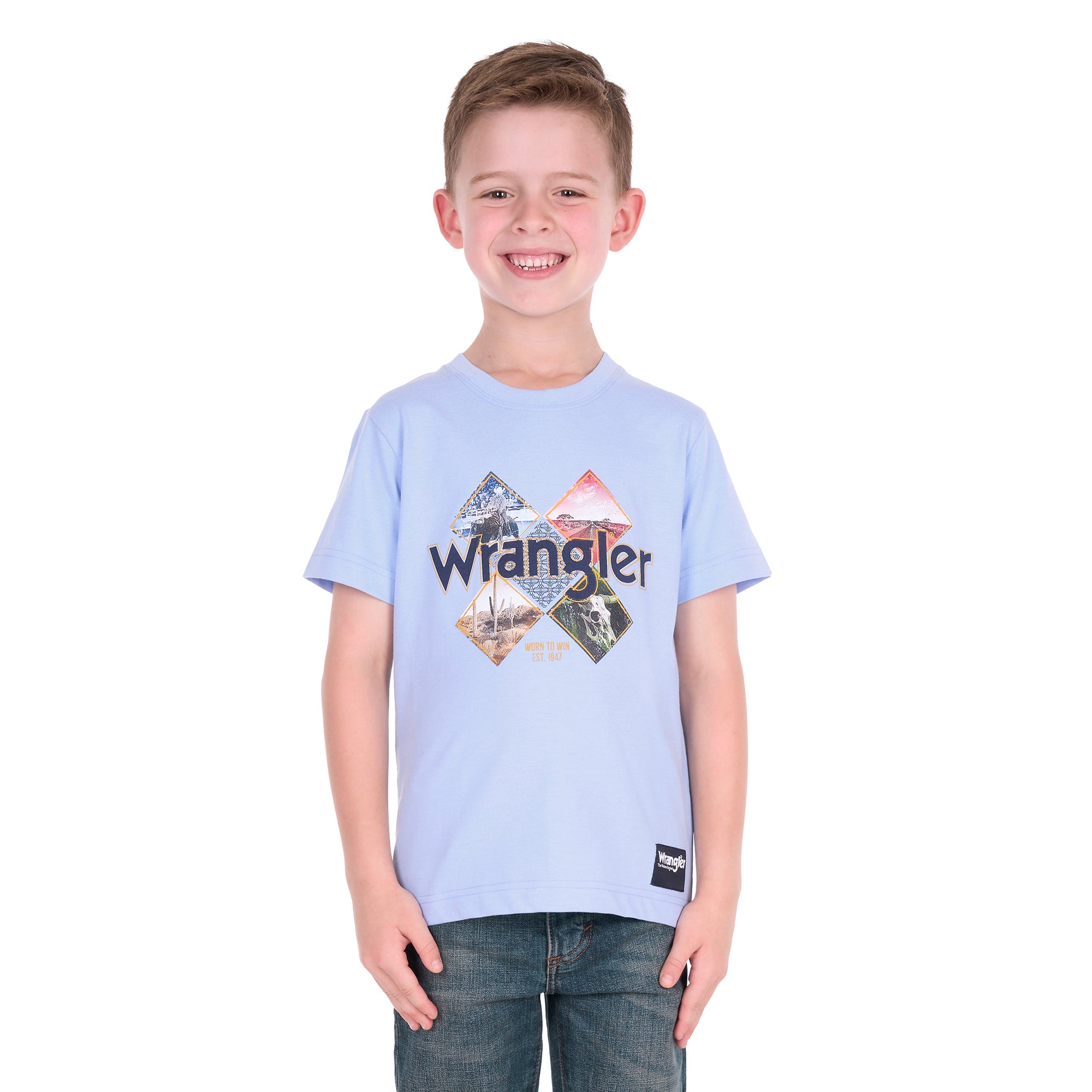 A smiling young boy wearing a light blue Wrangler Boys Mark Short Sleeve Tee (X6S3557688) with a graphic print on the front, paired with dark blue jeans, photographed against a solid white background.
