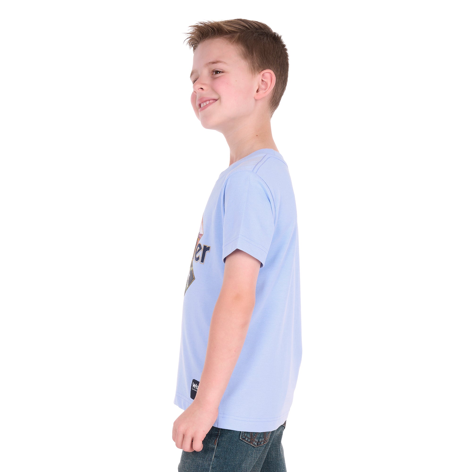 A smiling young boy in profile wearing a light blue Wrangler Boys Mark Short Sleeve Tee (X6S3557688) with a graphic print on the front, paired with dark blue jeans, photographed against a solid white background.