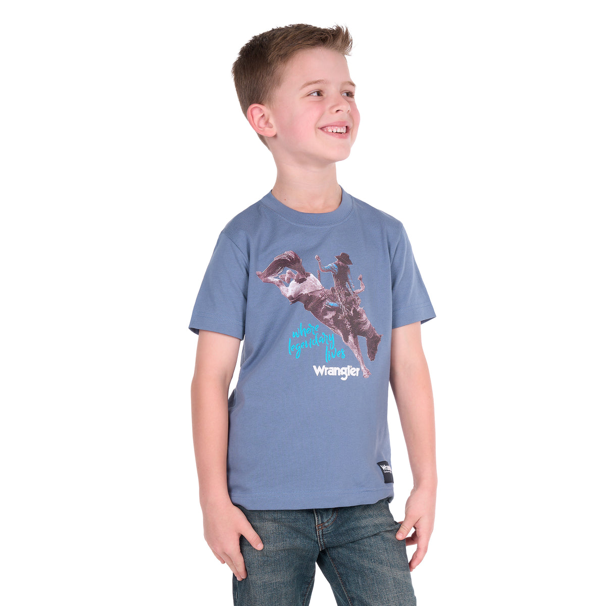 A young boy wearing a storm blue Wrangler Boys Michael Short Sleeve Tee (X6S3557684) with a cowboy graphic print, paired with blue jeans, standing against a solid white background.
