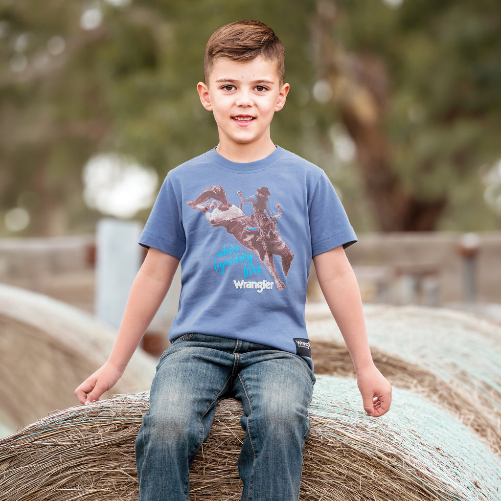 A young boy wearing a storm blue Wrangler Boys Michael Short Sleeve Tee (X6S3557684) with a cowboy graphic print, paired with blue jeans, sitting on a round hay bale outdoors.