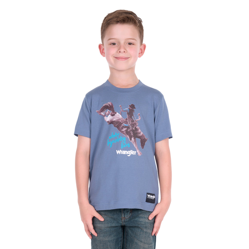 A young boy wearing a storm blue Wrangler Boys Michael Short Sleeve Tee (X6S3557684) with a cowboy graphic print, paired with blue jeans, standing against a solid white background.