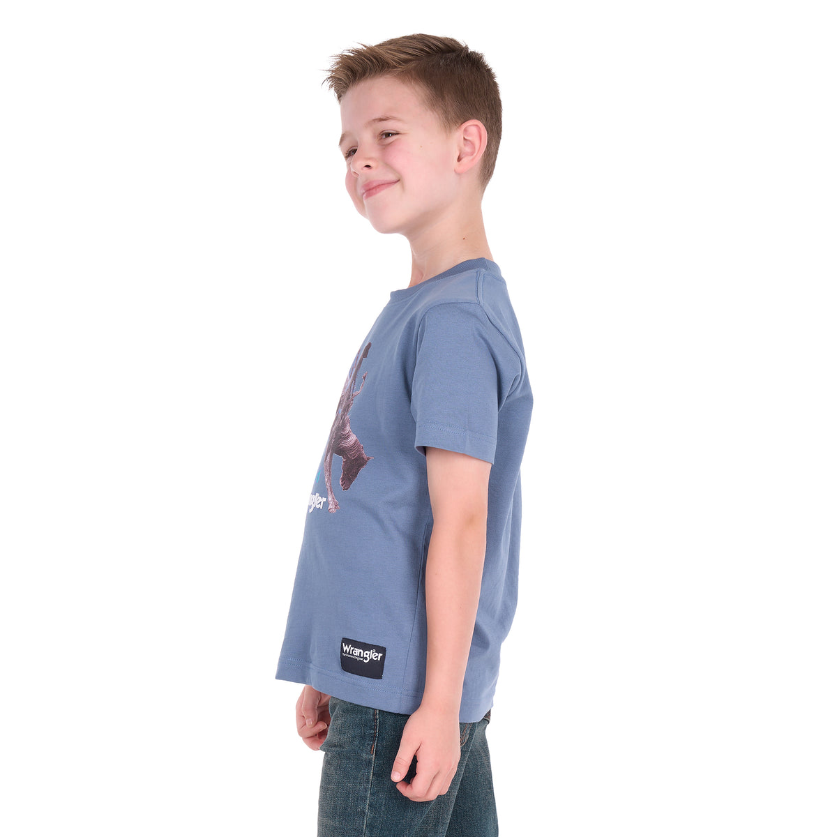A young boy in profile wearing a storm blue Wrangler Boys Michael Short Sleeve Tee (X6S3557684) with a cowboy graphic print, paired with blue jeans, standing against a solid white background.