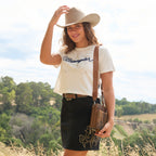 A woman wearing a Silverton cream Wrangler Brodie Hat (XCP1961HAT), a cream Wrangler Womens Cindy Crop Short Sleeve Tee (XCP2596647), a Wrangler Womens Rock Denim Skirt (X6S2409725) with gold embroidery, and a tan Wrangler Camile Wallet Bag (X6S2977BAG), standing outdoors in a grassy field with trees in the background.