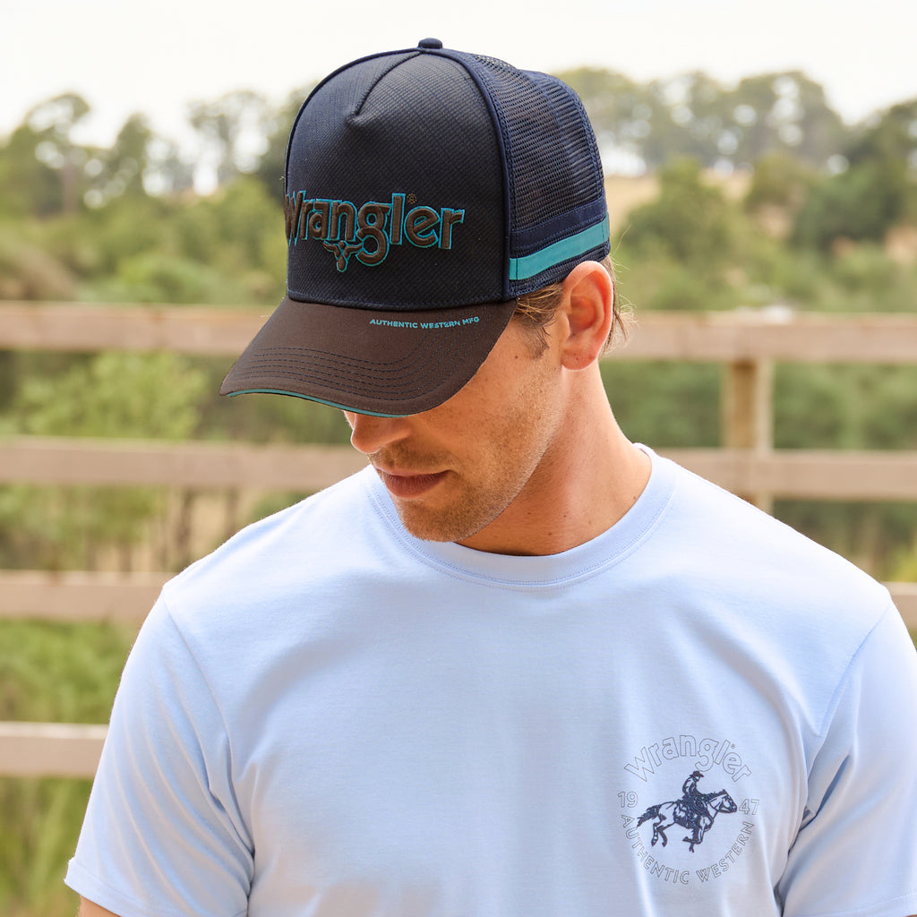 A man wearing a Wrangler Chase High Profile Trucker Cap (XCP1941CAP) in navy blue and tan, featuring an embroidered logo on the front, a brown visor with a turquoise trim, turquoise and navy side stripes and a navy mesh back, paired with a light blue Wrangler Steve T-shirt (X6S1557597), looking down in an outdoor setting with a wooden fence and trees in the background.