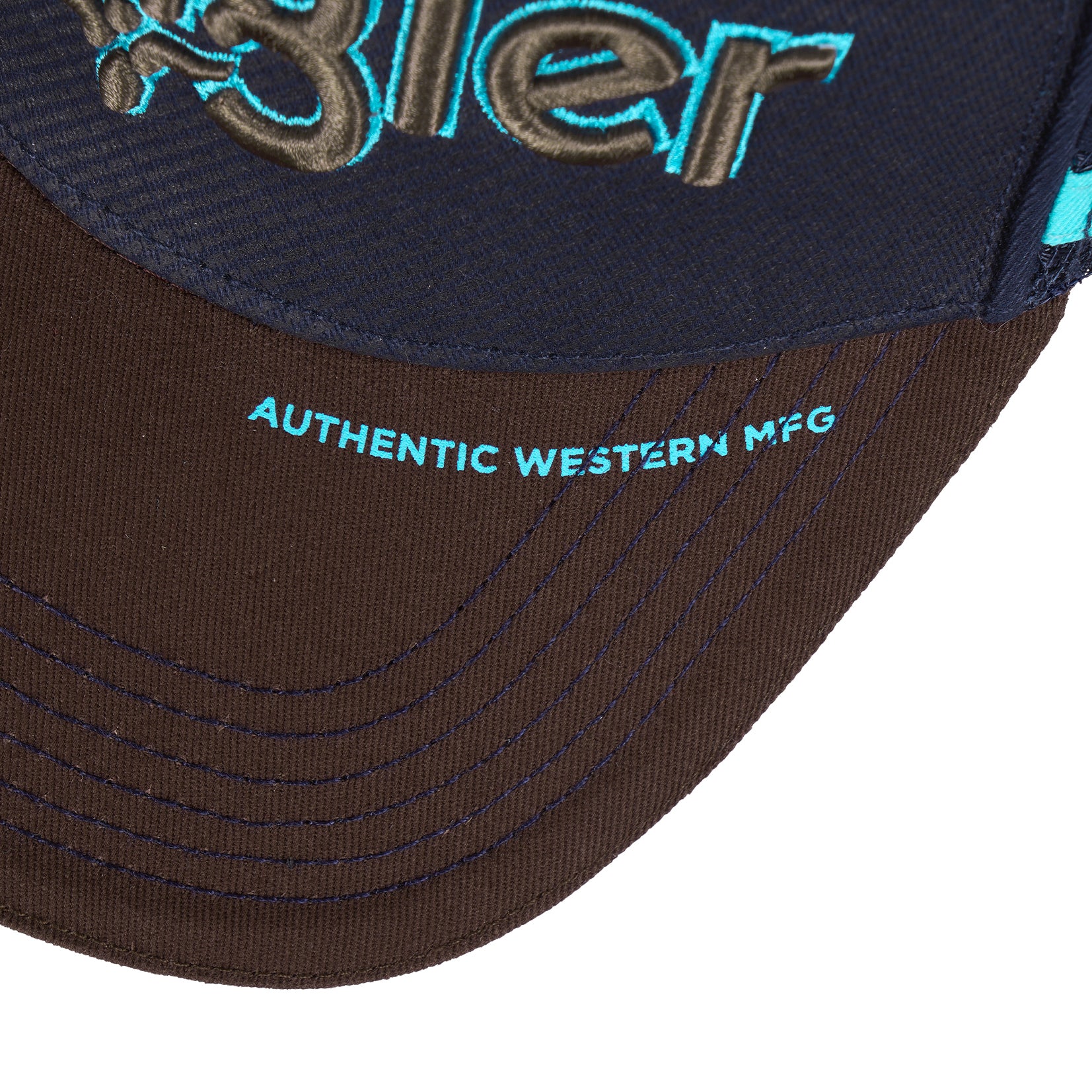 Close-up of a Wrangler Chase High Profile Trucker Cap (XCP1941CAP) in navy blue and tan, featuring an embroidered logo on the front, and a brown brim with 'AUTHENTIC WESTERN MFG' in turquoise on a white background.