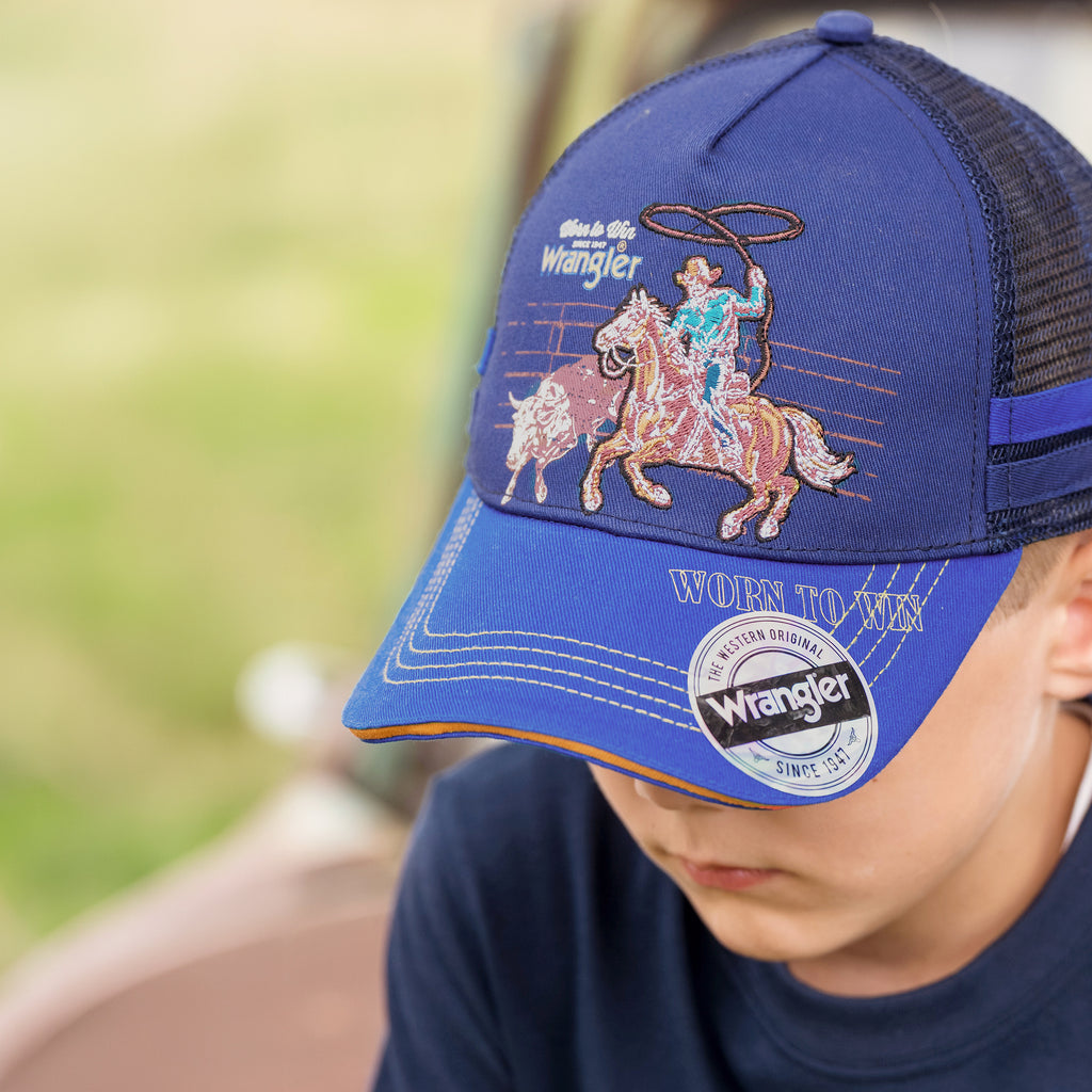 A close-up of a child wearing a navy/blue Wrangler Children's Abe Trucker Cap (X6S3946CAP) with a western cowboy embroidered on the front panel, a blue brim and navy mesh back panels, photographed outdoors with a blurred green background.