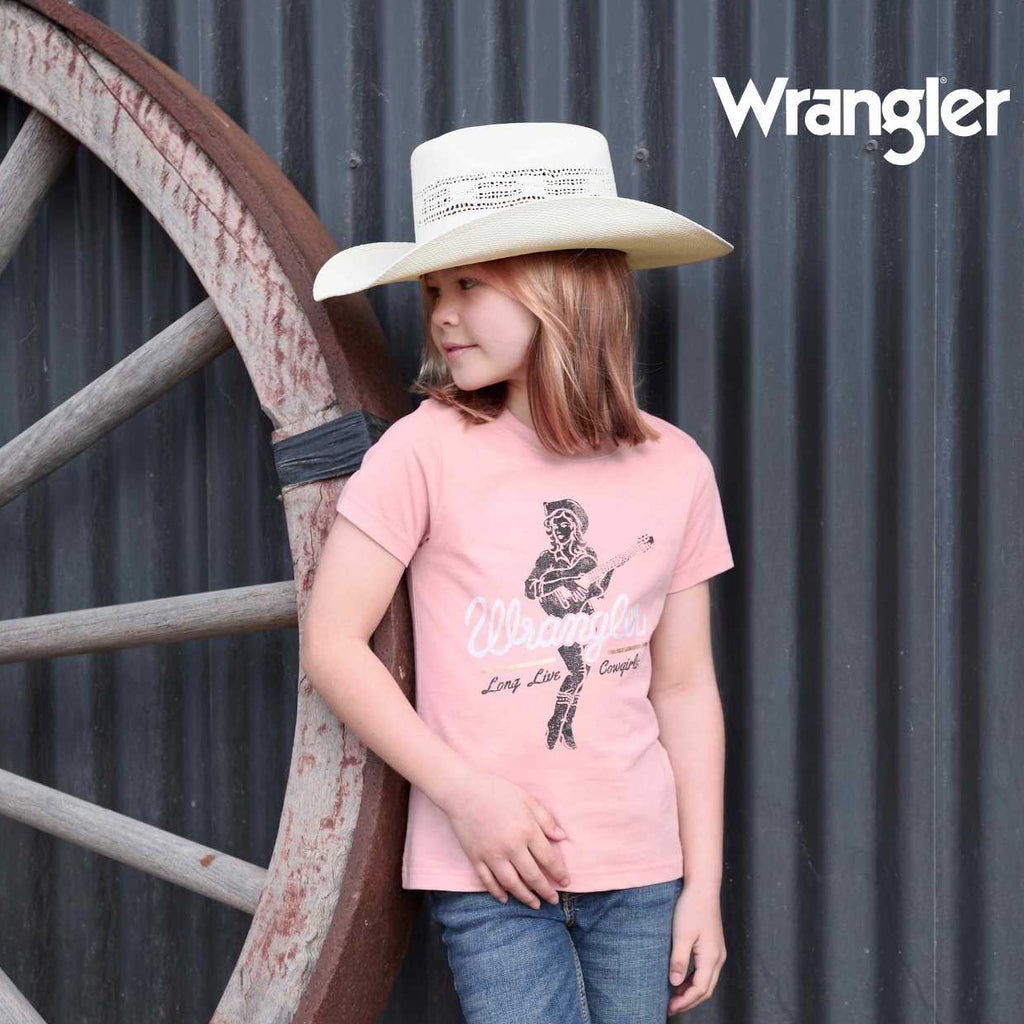 A young girl wearing a light pink Wrangler short-sleeve t-shirt with a western graphic, blue jeans, and a natural Wrangler Children's Boll Bangora Hat (X4Y3901BOL) posing next to a large rustic wooden wagon wheel in front of a corrugated metal wall. The Wrangler brand logo is visible in the top right corner.