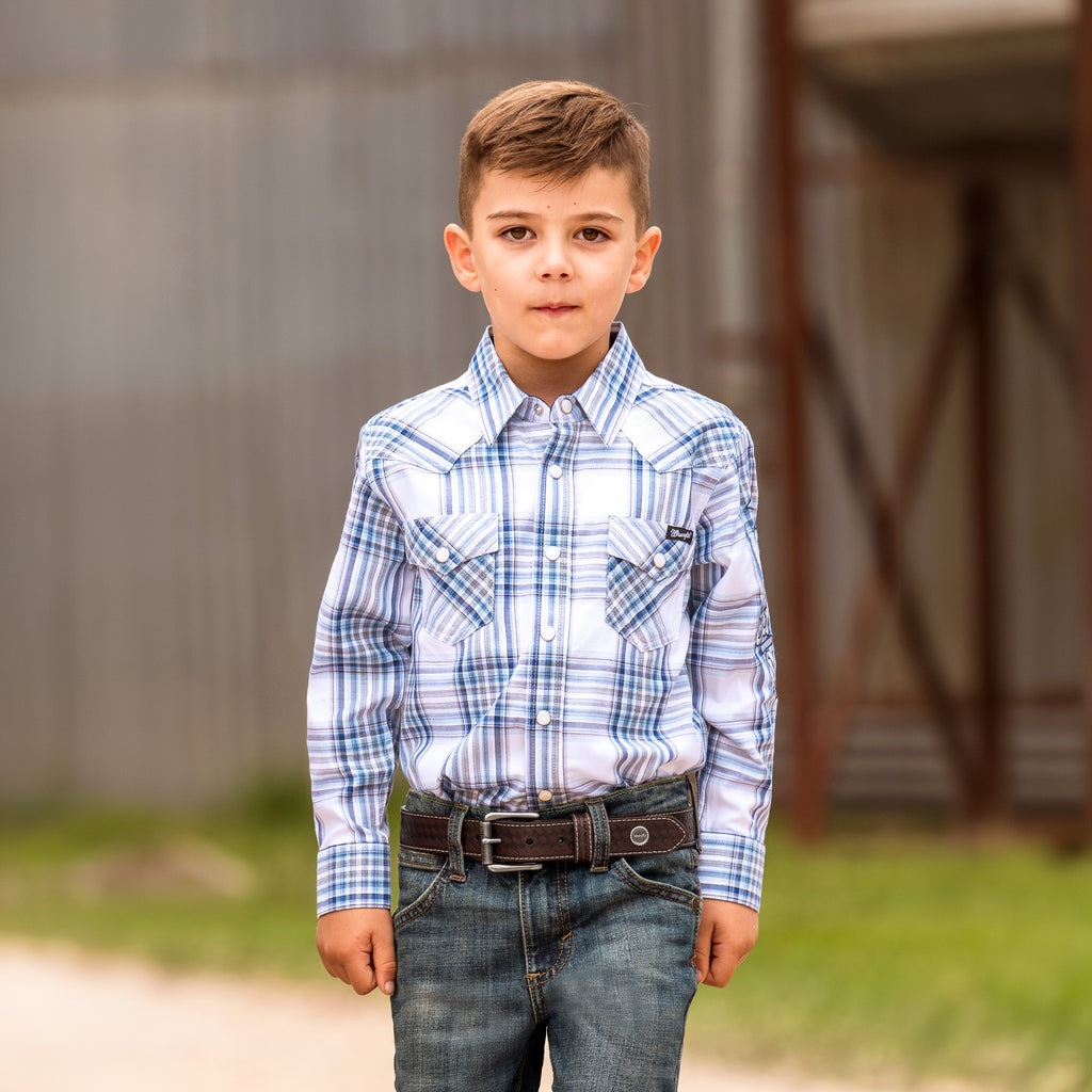 A boy modelling a brown woven dark tan Wrangler Children's Denny Belt (XCP7951BLT) with a silver-toned metal buckle and a circular silver Wrangler concho on the tip, in a rural setting.
