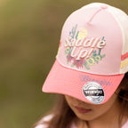 A close-up lifestyle shot of a child wearing a pink and white Wrangler Children's Imogen Full Back Cap (X6S5947CAP) with 'Saddle Up!' embroidered on the front, set against a soft, blurred outdoor background.