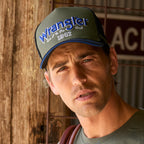 A man wearing a Wrangler Dave High Profile Trucker Cap (X6S1942CAP) with blue lettering on a dark green front panel, photographed against a rustic wooden wall.