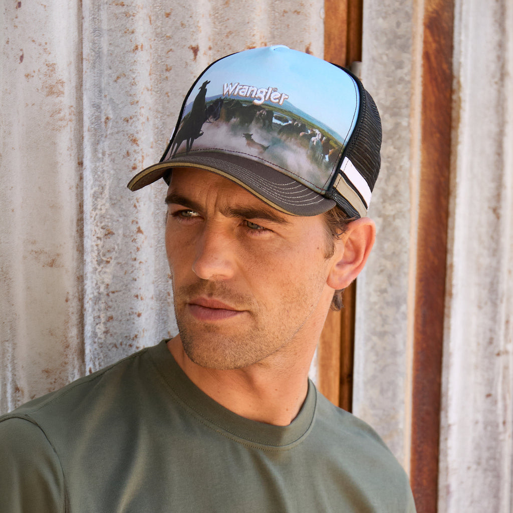 A man wearing a dark tan Wrangler Evan High Profile Trucker Cap (X6S1951CAP) featuring a brown brim, black mesh back, and a light blue printed front panel with a muster scene and an embroidered Wrangler logo, photographed against a rustic corrugated metal wall.