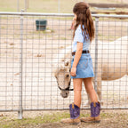 A young girl stands by a gate, looking at a white pony on the other side in a paddock. She is wearing a light blue t-shirt, a faded blue Wrangler Girls Grace Skirt (X6S5202720) with decorative stitching and purple and brown cowboy boots.