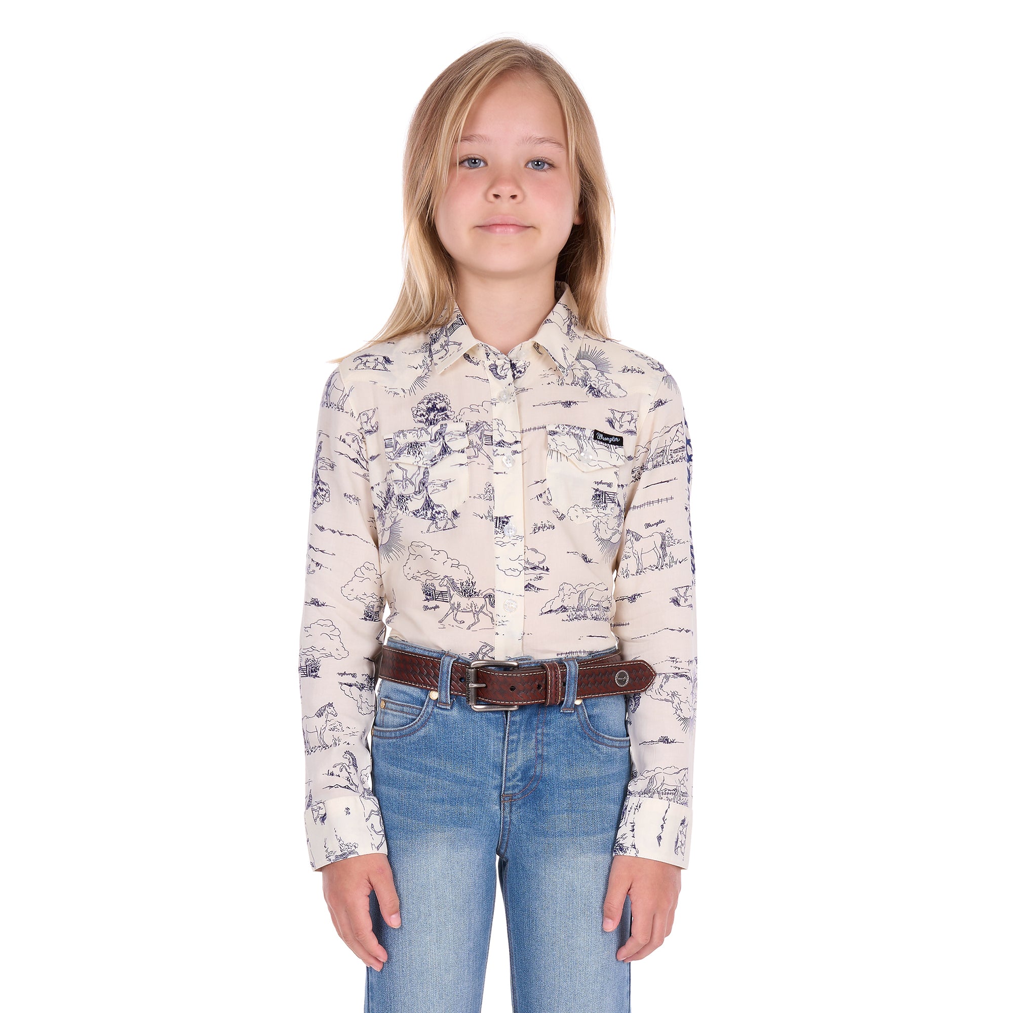 A young girl with blonde hair stands against a white background wearing a Wrangler Girls Ivory Logo Long Sleeve Shirt (X6S5127602) with a horse print on a cream base, paired with blue jeans and a dark tan Wrangler Children's Denny Belt (XCP7951BLT).