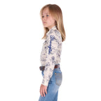 A young girl with blonde hair stands in profile against a white background wearing a Wrangler Girls Ivory Logo Long Sleeve Shirt (X6S5127602) with a horse print on a cream base, paired with blue jeans and a dark tan Wrangler Children's Denny Belt (XCP7951BLT).